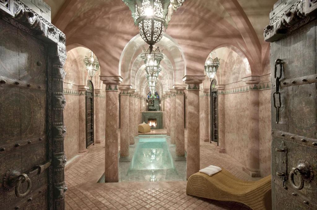 10 Unique Riads in Marrakech with Pool 42 Ornate, arched room with pink marble walls, columns, and lantern-style chandeliers—evoking the elegance of riads in Marrakech with pool. A small pool sits at the center, fireplace at the far end, and a woven lounge chair graces the tiled entrance.