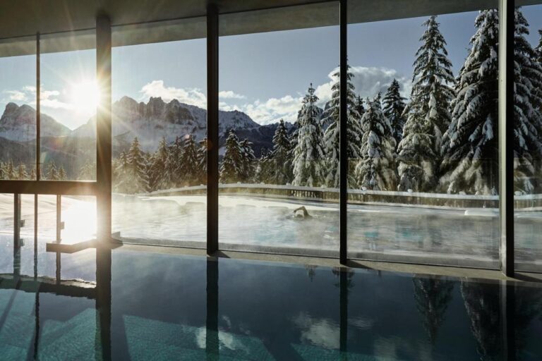 Indoor pool with large glass windows at one of the best hotels in the Dolomites overlooks snowy pine trees and mountain peaks, as sunlight streams in and creates a serene, warm atmosphere contrasting with the winter landscape outside.