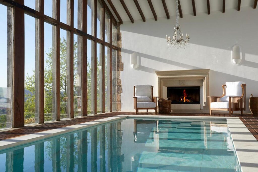 An indoor pool room with large floor-to-ceiling windows, two white armchairs by a lit fireplace, and a chandelier hanging from the beamed ceiling. Sunlight streams in, creating a relaxing atmosphere reminiscent of top hotels in Cantabria.