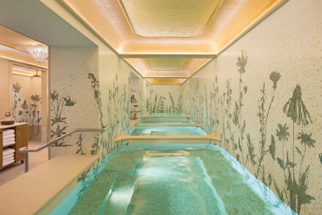 13 Unique Hotels in Paris for a Stylish Stay 7 A serene indoor spa in one of the unique hotels in Paris features a long pool with crystal-clear water, mosaic tile walls decorated with green botanical patterns, soft yellow ceiling lights, and a towel station on the left side.