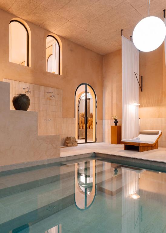 Top 12 Boutique Hotels in Porto, Portugal 33 A serene indoor pool in a modern, minimalist space—reminiscent of boutique hotels in Porto—with arched windows, soft beige walls, a lounge chair with sheer curtains, and a large round light fixture hanging from the ceiling.