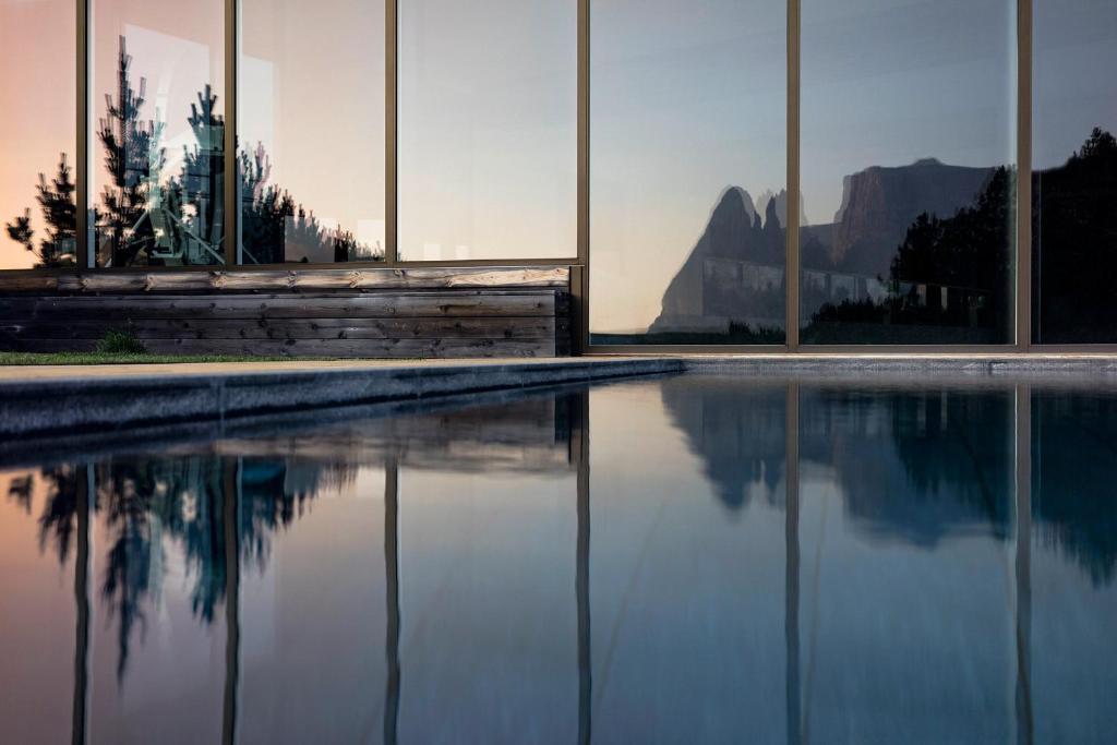 Large glass windows reflect a mountain landscape at sunset, with a pool in the foreground and pine trees and wooden planter boxes beside the building—capturing the elegance of the best hotels in the Dolomites.