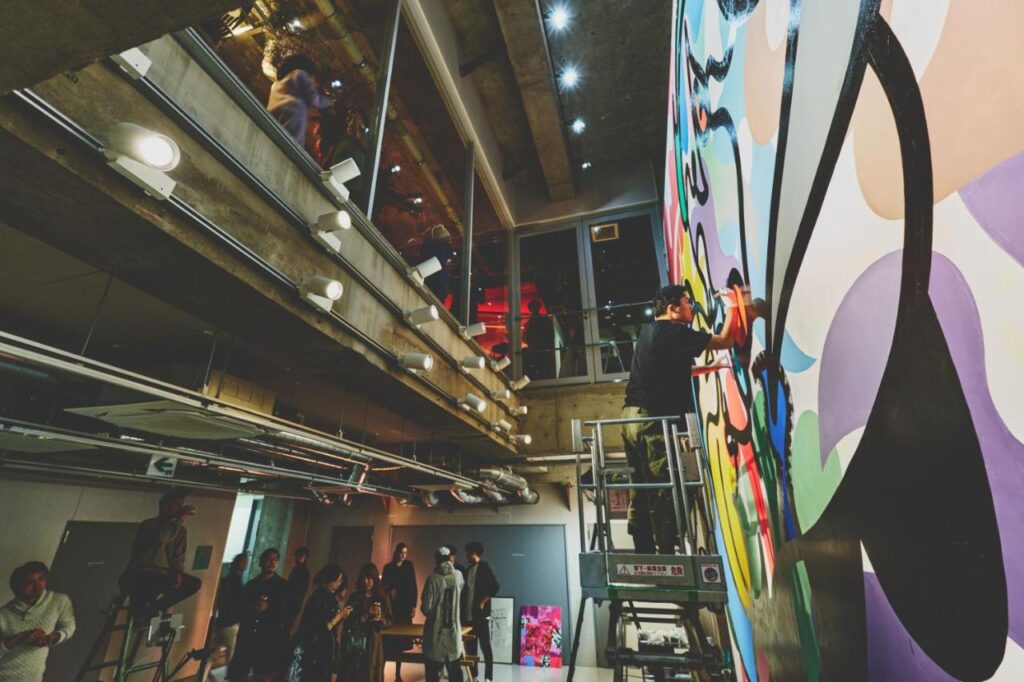 15 Unique Hotels in Tokyo: Coolest hotels 33 An artist stands on a ladder painting a colorful mural on a tall wall inside an industrial-style building—reminiscent of unique hotels in Tokyo—while several people watch from below and an upper balcony.