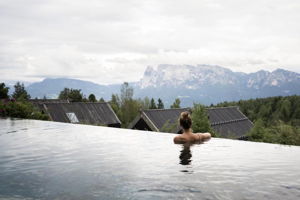 A person with their hair in a bun relaxes in an infinity pool at one of the best hotels in the Dolomites, overlooking rustic rooftops, dense green trees, and distant mountains under a cloudy sky.