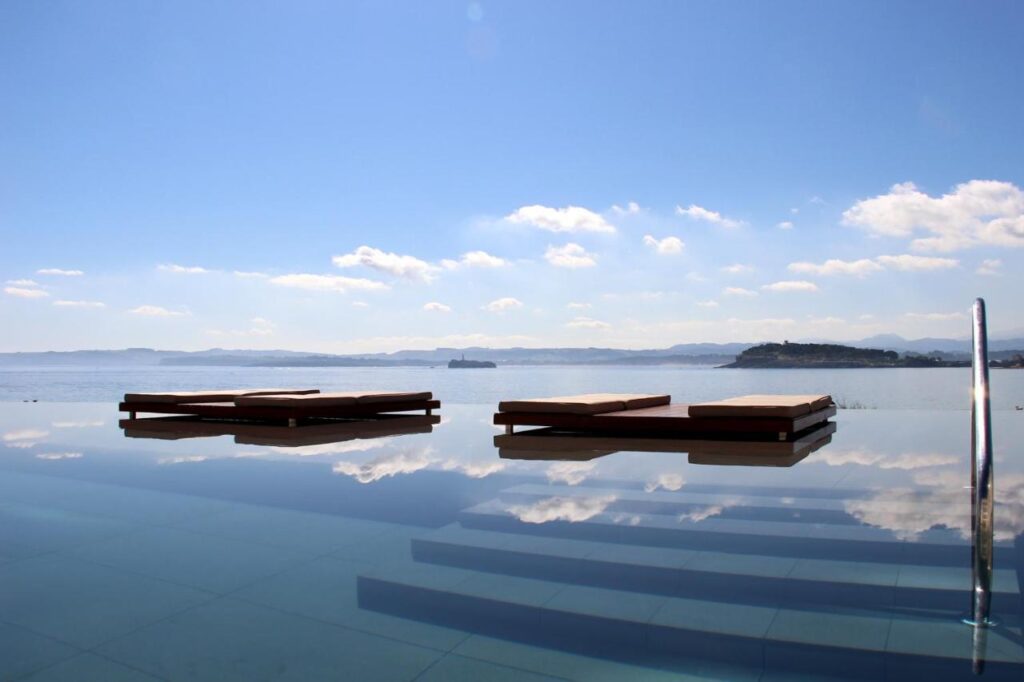 Two wooden lounge chairs sit on the edge of an infinity pool at one of the tranquil hotels in Cantabria, overlooking a calm, expansive body of water and distant hills beneath a clear sky with scattered clouds.
