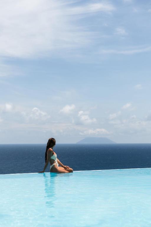 7 Best Hotels in the Aeolian Islands 17 A woman in a swimsuit sits at the edge of an infinity pool, overlooking a calm blue sea and a distant island—an idyllic moment reminiscent of luxury hotels in the Aeolian Islands, with a partly cloudy sky completing the serene scene.