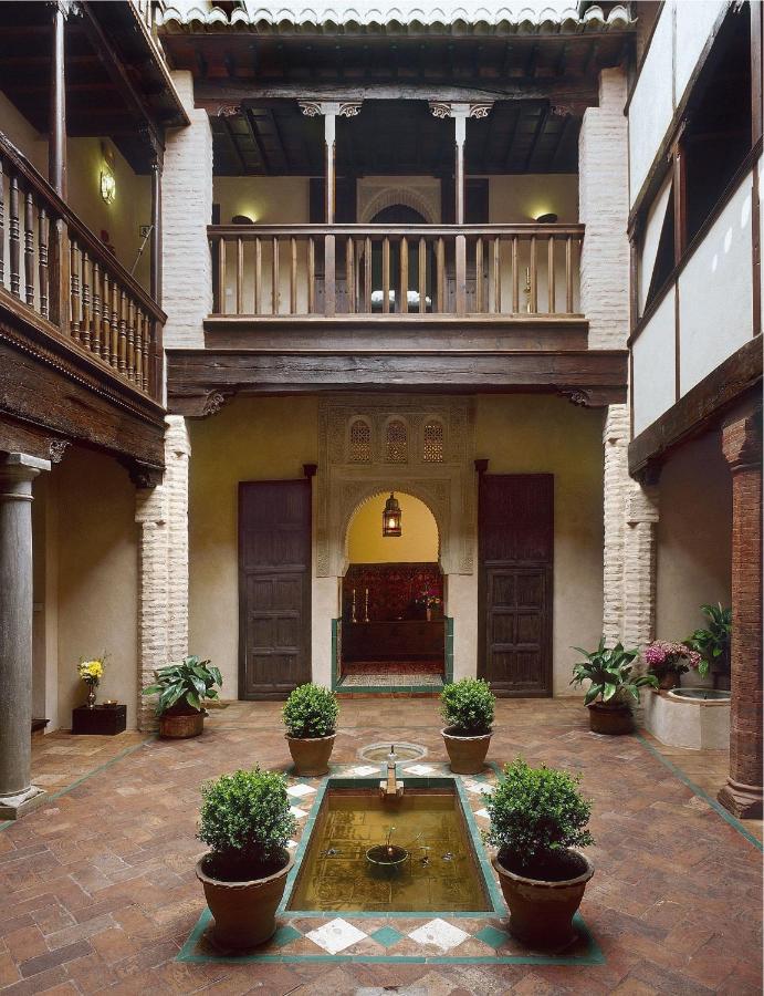 A traditional courtyard like those found in boutique hotels in Granada, with terracotta flooring, potted plants, a central rectangular fountain, wooden balcony, arched doorways, tiled roof, columns, and warm lighting.