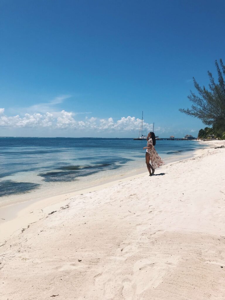 Top Hotels in Tulum, Mexico: Luxury and Beachfront Stays 2 A woman in a white dress stands on a sandy beach, looking out at the calm blue sea near some of the top hotels in Tulum. A sailboat is anchored in the distance while trees line the far edge of the shore.