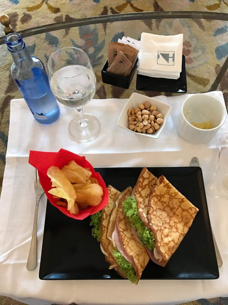 A plate with two folded crepe sandwiches filled with ham and lettuce, served with potato chips in a red napkin—reminiscent of light bites often enjoyed at hotels in Cantabria. Nearby are water, pistachios, and sugar packets on a white tablecloth.