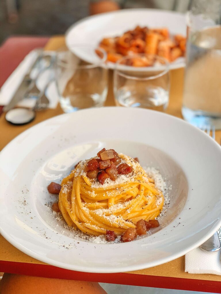 13 UNIQUE Boutique Hotels in Rome 2 A plate of spaghetti carbonara topped with pancetta and grated cheese is in focus on a table at one of the boutique hotels in Rome, with glasses, cutlery, and another pasta dish blurred in the background.
