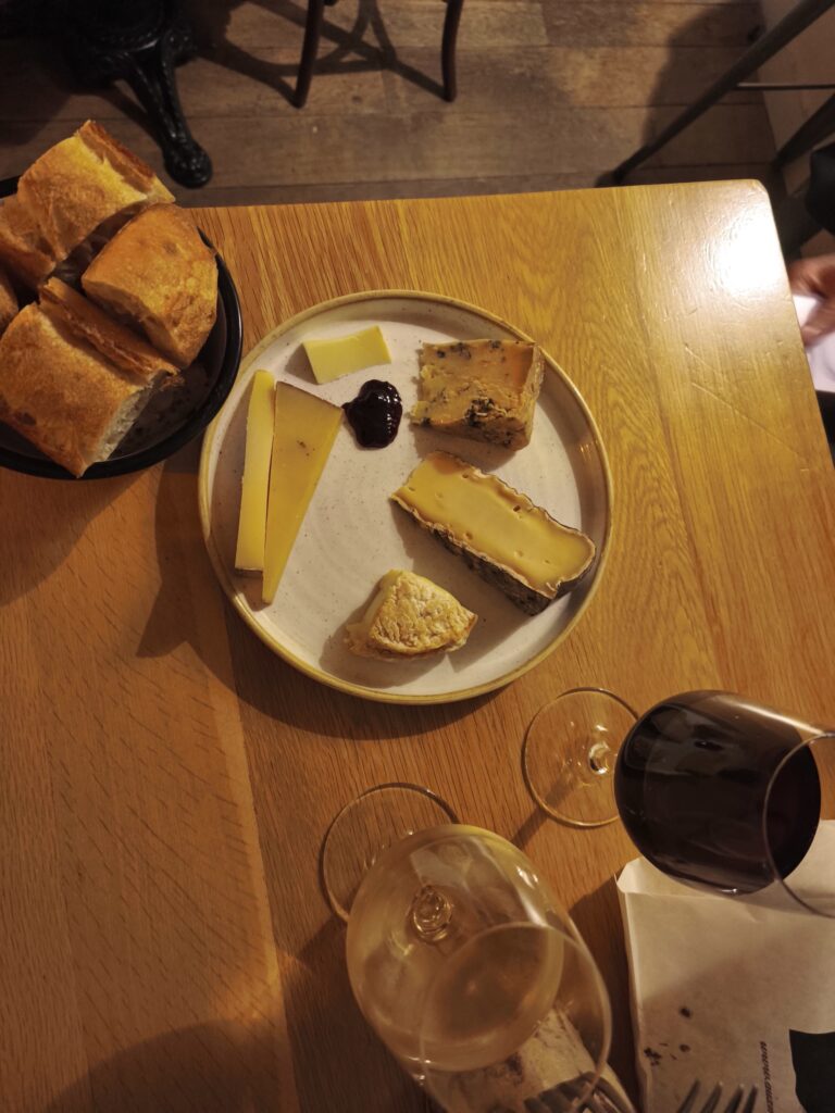 13 Unique Hotels in Paris for a Stylish Stay 2 A plate with assorted cheeses and a dollop of jam sits on a wooden table, alongside bread and wine—just the kind of spread you might find in one of the unique hotels in Paris. A fork and napkin are also visible.