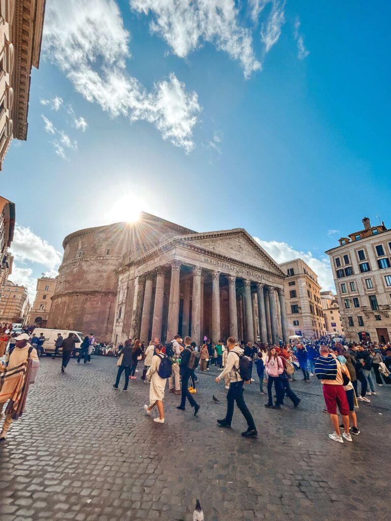 13 UNIQUE Boutique Hotels in Rome 56 Crowds of people gather in front of the Pantheon in Rome on a sunny day, sunlight shining over the historic columns and a vibrant blue sky with scattered clouds—steps away from some of the finest boutique hotels in Rome.