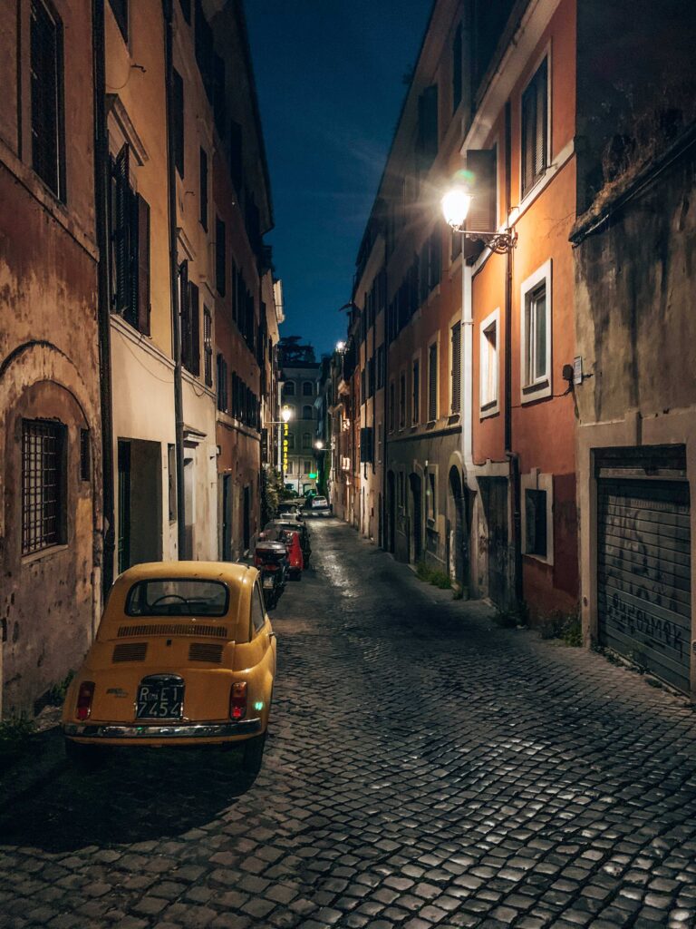 13 UNIQUE Boutique Hotels in Rome 59 A narrow cobblestone street in a European city at night, lined with tall, old buildings—much like the charming lanes near boutique hotels in Rome. A vintage yellow Fiat is parked in the foreground beneath a streetlamp, others line the street.
