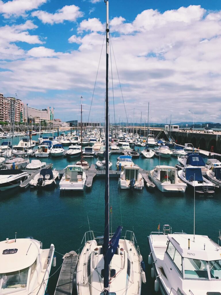A marina filled with numerous docked boats and yachts under a partly cloudy blue sky, with apartment buildings and hotels in Cantabria visible on the left, overlooking calm turquoise water in the harbor.