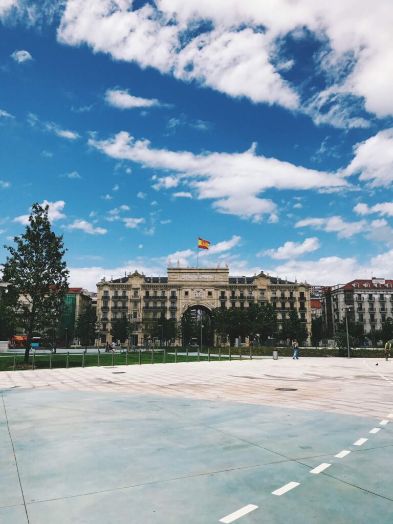 A large historic building with a Spanish flag on top stands under a bright blue sky with scattered clouds. The foreground features an open plaza with a few trees and people walking, reminiscent of the grand hotels in Cantabria.