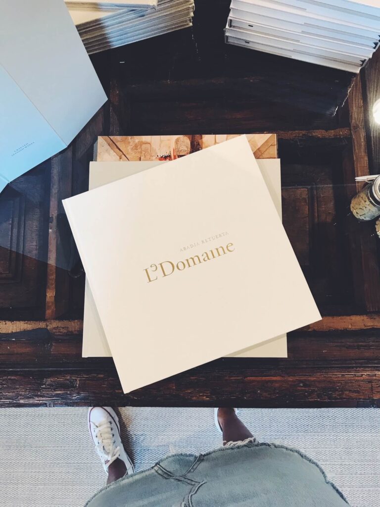 4 Best Hotels in Ribera del Duero, Spain 9 A person in ripped jeans and white sneakers stands by a coffee table with a large white book titled L’Domaine in gold letters—a nod to the refined hotels in Ribera del Duero—while other books and papers are stacked around it.