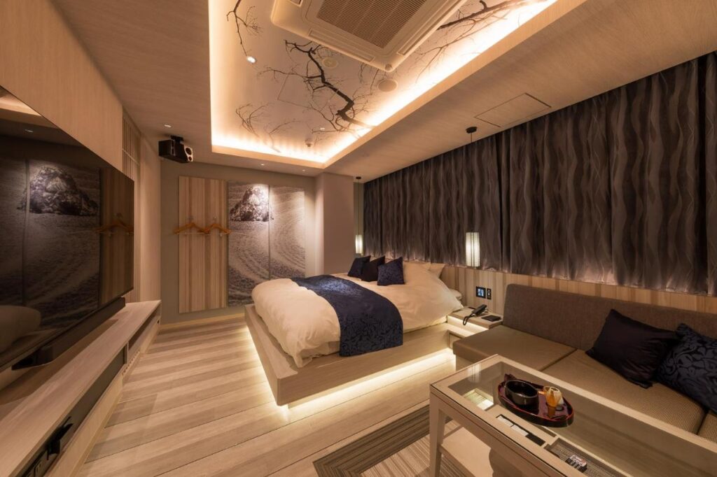 15 Unique Hotels in Tokyo: Coolest hotels 35 A modern hotel room with a large bed, dark blue accents, decorative ceiling art, and a wall-mounted TV. Enjoy a cozy, elegant atmosphere in one of the unique hotels in Tokyo, complete with a sofa and glass coffee table with a tea set.