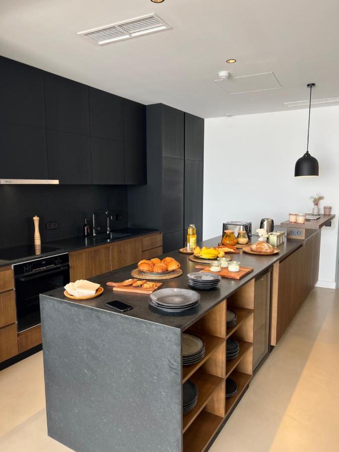 Unique Boutique Hotels in Menorca 33 Modern kitchen with black and wood cabinets, a dark stone island featuring stacked plates, and a breakfast spread of croissants and fruit—reminiscent of the inviting ambiance found in boutique hotels in Menorca.
