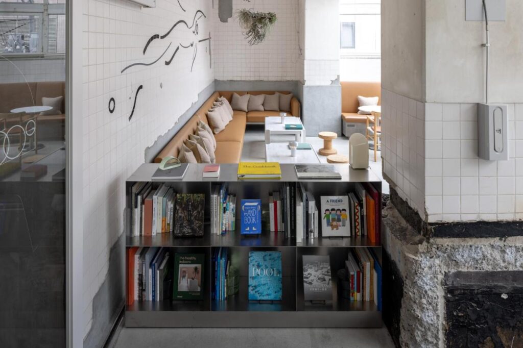 7 Best Hotels in Hiroshima, Japan 4 A modern, minimalist lounge in the style of the best hotels in Hiroshima, Japan, featuring a low bookshelf filled with books, beige cushioned seating, white tables, wall art, and hanging plants in a bright space with tiled walls.