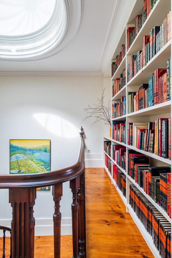 Top 12 Boutique Hotels in Porto, Portugal 39 A bright hallway with a large skylight, wooden handrail, built-in bookshelves filled with books, a framed landscape painting on the wall, and a bare branch in a vase—reminiscent of boutique hotels in Porto.