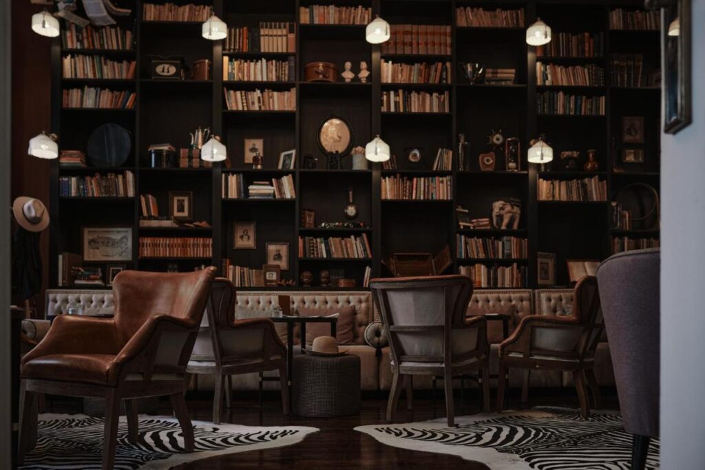 Top 12 Boutique Hotels in Porto, Portugal 10 A cozy library lounge found in boutique hotels in Porto, with leather and upholstered chairs, round tables, and a large bookshelf filled with books and vintage decor. Soft pendant lights hang overhead, while zebra-print rugs warm the wooden floors.