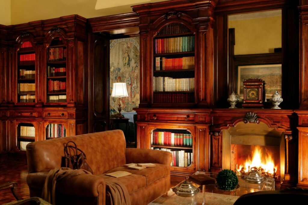 13 UNIQUE Boutique Hotels in Rome 21 A cozy library inspired by boutique hotels in Rome, Italy, with wooden bookshelves filled with books, a lit fireplace, a brown leather sofa, a small table with items, and warm ambient lighting creating an inviting atmosphere.