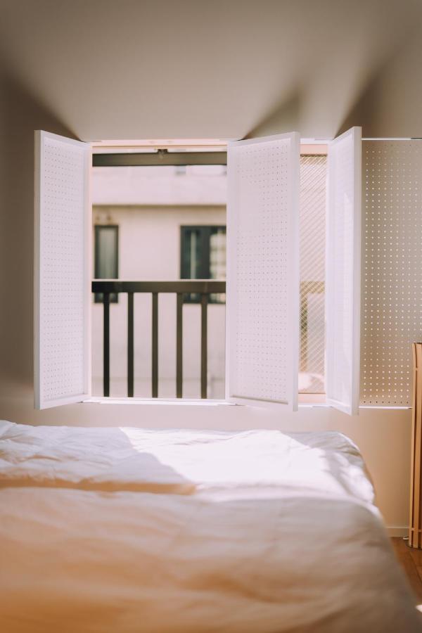15 Unique Hotels in Tokyo: Coolest hotels 56 A sunlit bedroom in one of the unique hotels in Tokyo, with white bedding, an open window with white shutters, and a view of a building and balcony railing outside. Soft light casts gentle shadows on the bed.