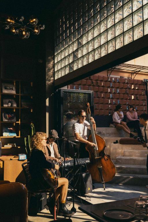 A small jazz band performs indoors by large windows in one of the boutique hotels in Athens; one person plays guitar and sings, another plays double bass, and a third plays keyboard as sunlight streams in and several people watch from outside on tiered steps.