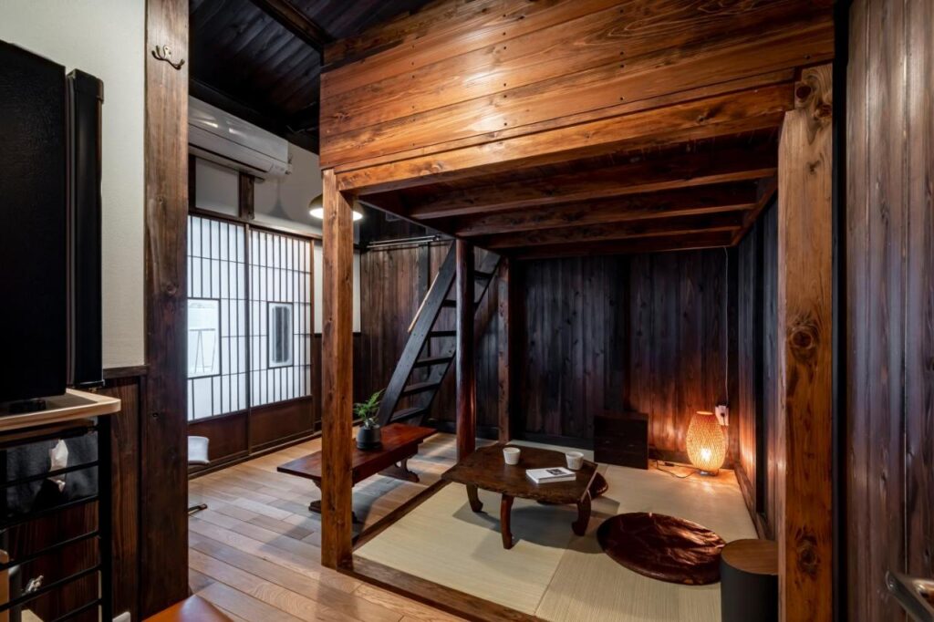 15 Unique Hotels in Tokyo: Coolest hotels 20 A cozy Japanese-style living room, inspired by unique hotels in Tokyo, features dark wooden walls and beams, a loft with stairs, a low table with floor cushions, a tatami mat, soft lighting, and a shoji screen window.
