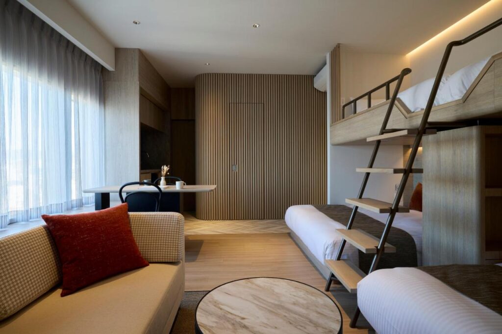 7 Best Hotels in Hiroshima, Japan 7 Modern hotel room in one of the best hotels in Hiroshima, Japan, featuring a bunk bed, sofa with red cushion, round marble coffee table, desk with two chairs by a window with sheer curtains, and minimalist wooden décor.