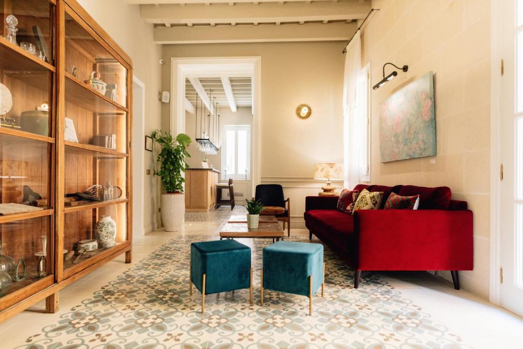 Unique Boutique Hotels in Menorca 48 Bright living room with a red velvet sofa, two teal ottomans, patterned tile floor, wooden display cabinet, wall art, and a view into a kitchen area—evoking the chic style found in boutique hotels in Menorca. Natural light pours in from large windows.