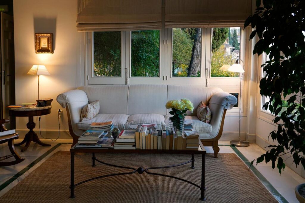 A cozy living room with a white sofa, pillows, and a coffee table covered with books and flowers, echoing the inviting charm of boutique hotels in Granada. Large windows let in natural light and reveal trees outside, while lamps and a plant add warmth to the space.