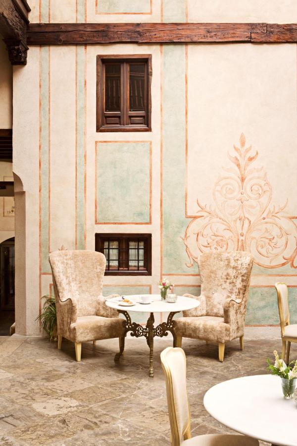 Elegant seating area with two ornate, high-backed chairs and a round white table set for tea and pastries, evoking the charm of boutique hotels in Granada against a decorative pastel wall with wooden windows in a bright, rustic space.