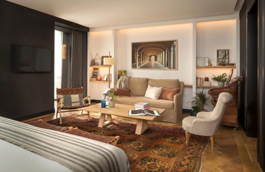 13 Unique Hotels in Barcelona, Spain 9 A cozy living area with a tan sofa, armchair, and wooden coffee table on a patterned rug—much like those found in unique hotels in Barcelona—decorated with books, plants, art on the walls, warm lighting, and modern decor.