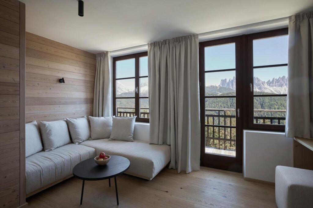 A modern, cozy living room with a light-colored L-shaped sofa, round black coffee table with apples, wooden walls and flooring, large windows with curtains, and a mountain view reminiscent of the best hotels in the Dolomites.