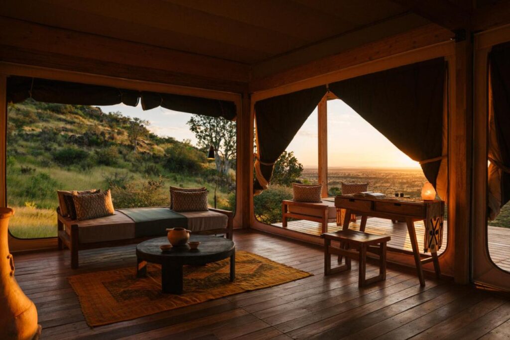 Unique Safari Lodges in Namibia: Wildlife and Adventure 2 A cozy, open-air lounge with wooden floors and rustic furniture overlooks a sunlit savanna at sunset—an inviting retreat inspired by the charm of safari lodges in Namibia, blending indoor comfort with sweeping views through large open windows.
