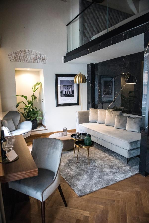 13 UNIQUE Boutique Hotels in Rome 15 Modern living room with a light gray sofa, matching armchairs, a gold floor lamp, houseplants, and framed artwork—echoing the chic style of boutique hotels in Rome, Italy. A glass balcony overlooks this cozy and stylish space.