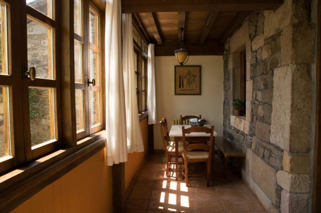 A sunlit, rustic dining area with wooden windows, a table set for two with wooden chairs, stone walls, white curtains, and a potted plant on the windowsill—just like those charming hotels in Cantabria. Warm natural light fills the cozy space.