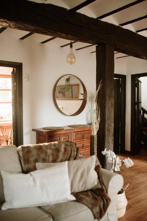 A cozy living room, reminiscent of charming hotels in Cantabria, with a beige sofa, white and brown pillows, a wooden dresser topped by a round mirror, exposed beams, and natural light streaming through nearby doors.