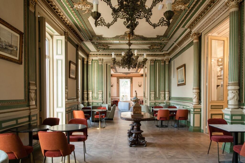 Elegant, ornate room in one of the finest hotels in Cantabria, with green and gold wall moldings, high decorated ceiling, chandeliers, tall windows, round tables, and red velvet chairs neatly arranged as natural light fills the space.