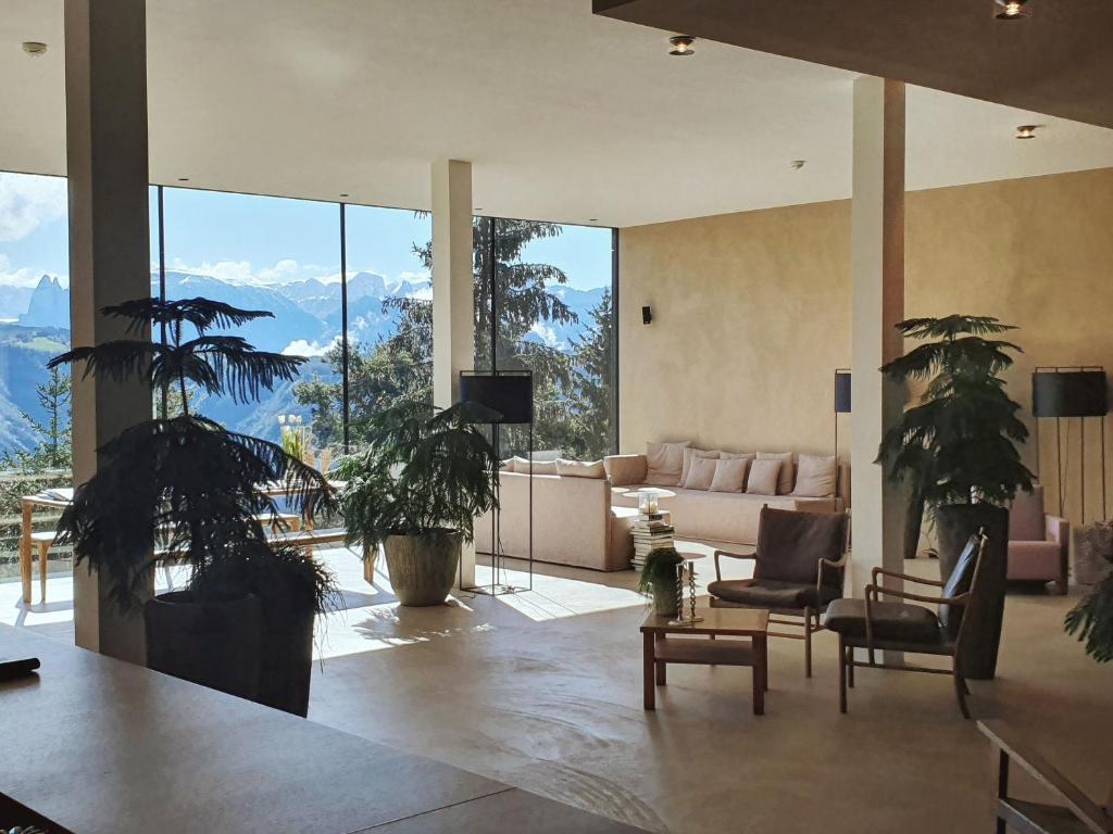 A modern living room with large windows, potted plants, neutral-toned furniture, and a view of mountains and trees—reminiscent of the serene vistas found at the best hotels in the Dolomites under a bright sky.