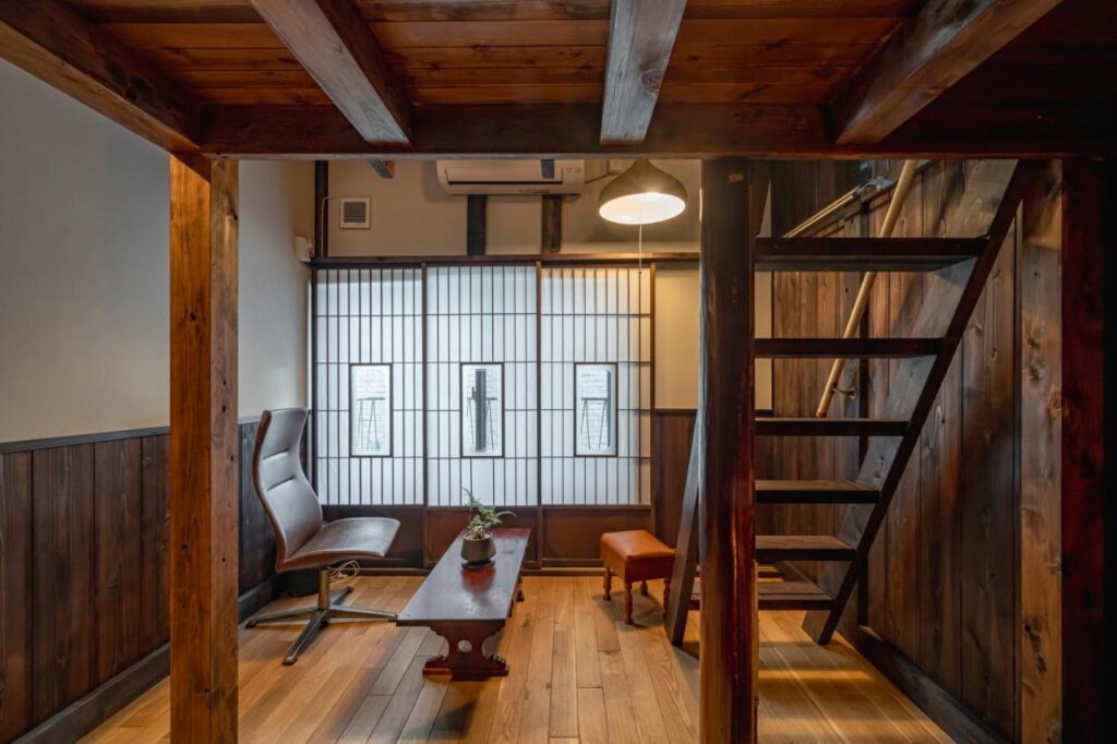 15 Unique Hotels in Tokyo: Coolest hotels 19 A cozy room with wooden beams and stairs, a tall window with shoji-style screens, a modern chair and ottoman, and a small wooden table with a plant—this inviting space reflects the charm found in unique hotels in Tokyo.