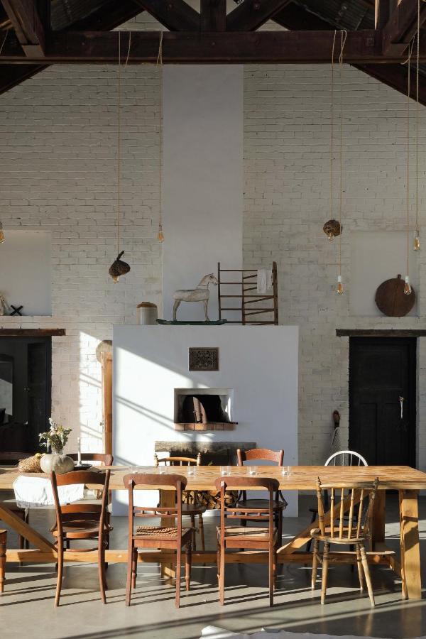 Unique Places to Stay in Cape Town 22 A rustic dining room with mismatched wooden chairs around a wooden table, a white brick fireplace, high ceilings with exposed beams, and sunlight streaming in—perfect for those seeking unique places to stay in Cape Town.