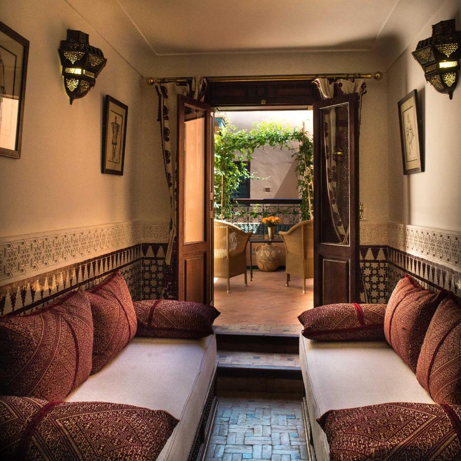 10 Unique Riads in Marrakech with Pool 6 Cozy Moroccan-style room with patterned cushions on two sofas, intricate wall tiles, and lamps—reminiscent of riads in Marrakech with pool—leading to an outdoor patio with a table, chairs, plants, and sunlight filtering in through double doors.