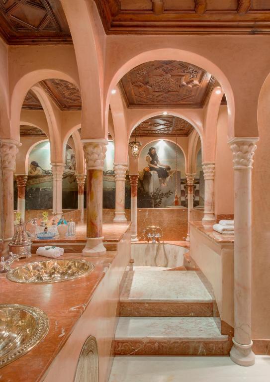 10 Unique Riads in Marrakech with Pool 41 Luxurious bathroom inspired by riads in Marrakech with pool, featuring marble steps to a sunken tub, gold sinks, ornate arches, carved columns, and a mural. Warm lighting enhances the elegant, classical decor.