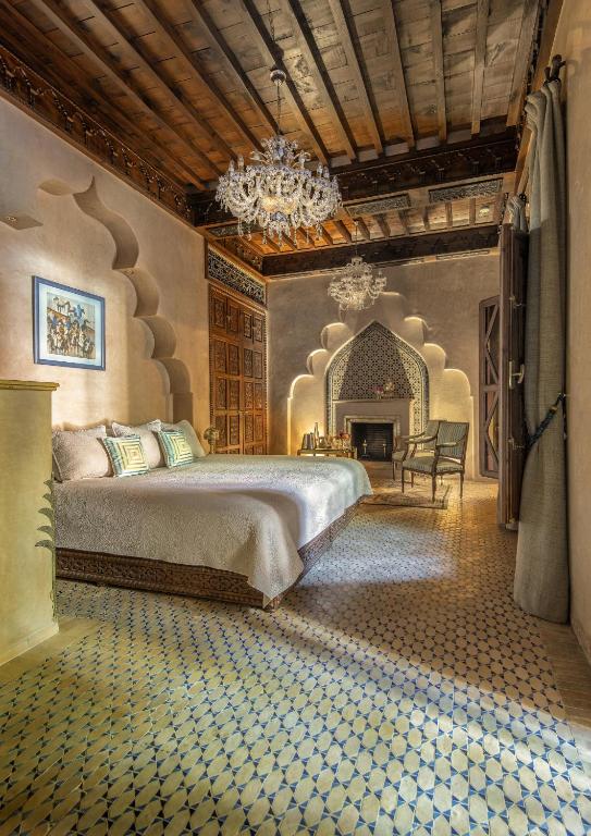 10 Unique Riads in Marrakech with Pool 39 A luxurious bedroom inspired by riads in Marrakech with pool, featuring Moroccan-style decor, intricate tile flooring, arched niches, chandeliers, a large bed with white linens, ornate wood ceiling, and a cozy seating area near a fireplace.