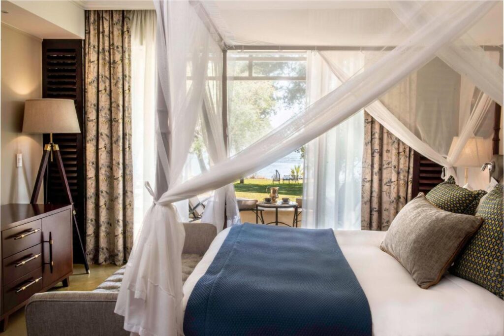 Best Hotels in Victoria Falls: Unique Luxury & Safari Stays 4 A cozy, elegant bedroom—reminiscent of the best hotels in Victoria Falls—features a canopy bed draped in white fabric, blue blanket, decorative pillows, cushioned bench, side table, lamp, floral curtains, and garden and water views through large windows.