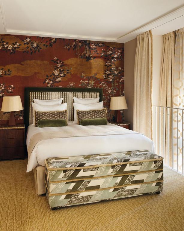 13 Unique Hotels in Paris for a Stylish Stay 34 A cozy bedroom in one of the unique hotels in Paris, featuring a double bed with a patterned headboard, floral and gold wallpaper, matching pillows, bedside tables with lamps, beige curtains, and a patterned bench at the foot of the bed.