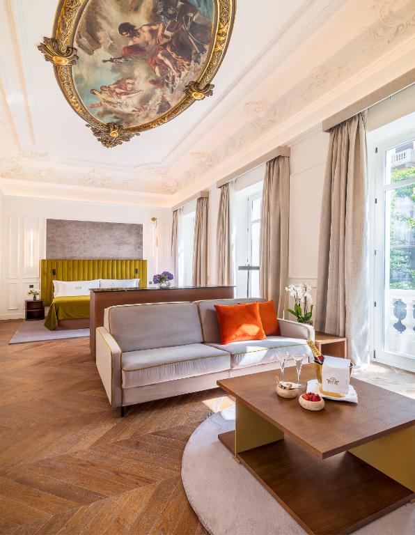 Elegant hotel suite with a bed, sofa, and coffee table on a wooden floor. Large windows with gray curtains let in natural light. The ornate ceiling with a painted mural captures the charm of boutique hotels in Granada.
