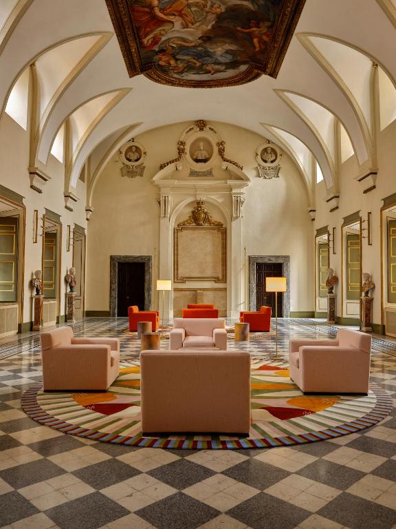 13 UNIQUE Boutique Hotels in Rome 48 Spacious ornate hall with vaulted ceiling, classical statues, and a large colorful round rug surrounded by modern pastel armchairs and sofas—evoking the elegance of boutique hotels in Rome. A painted ceiling and tall lamps complete the historic atmosphere.